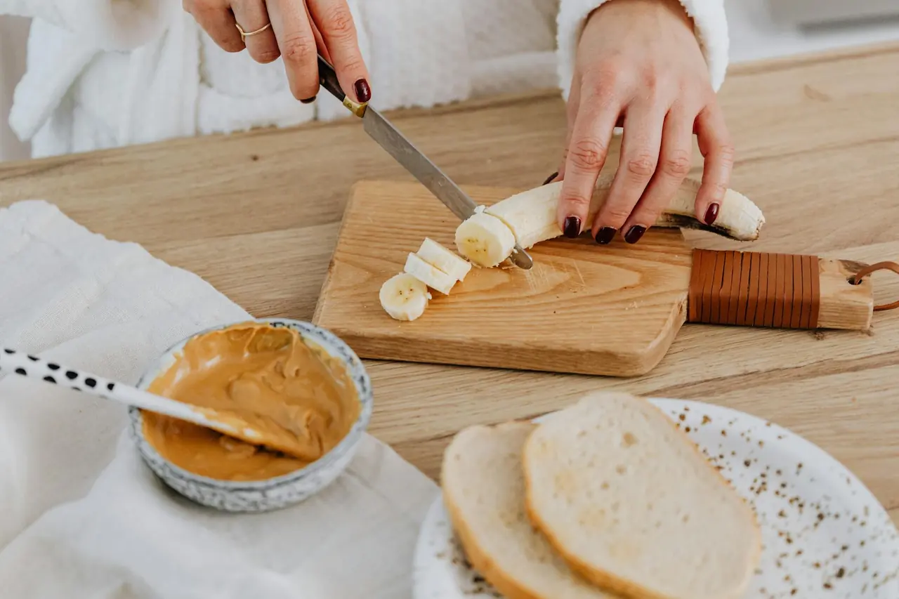Hand slicing banana with bread and peanut butter on a wooden board.