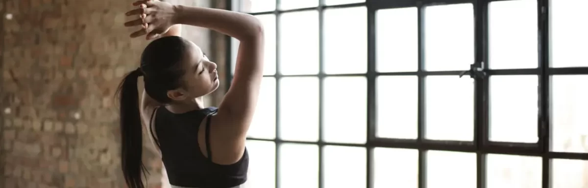 Athletic woman stretching her arms indoors beside a large window after exercise.