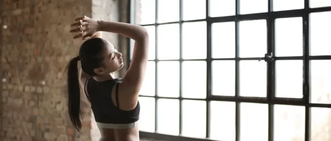Athletic woman stretching her arms indoors beside a large window after exercise.