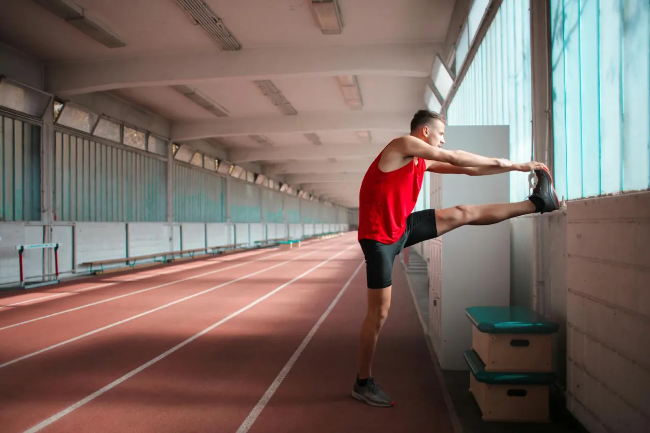 Male athlete stretching indoors on a sunny track, representing warm-up and recovery work.
