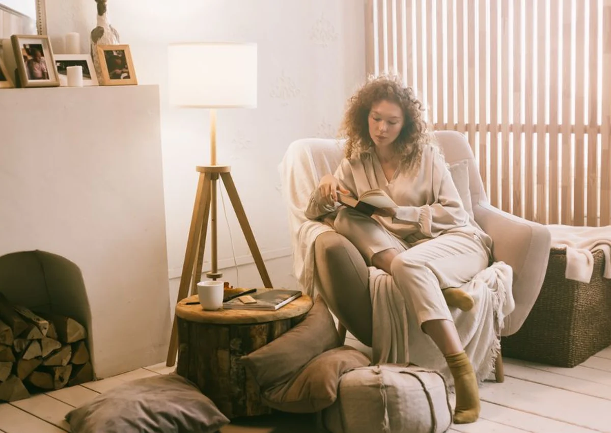 A person reading in an armchair beside a lamp and small table.